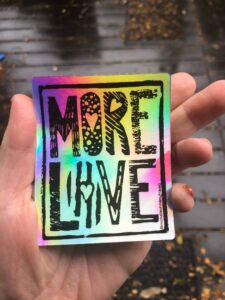 Michelle, a white non-binary person, holds the sticker in their left hand. A thumb is visible on the left side of the sticker. The four fingers are on the right side of the sticker. The edge of Michelle's shortened pinkie is visible holding the edge of the sticker. Horizontal brown wood decking is wet with leaves on it blurry behind the hand.