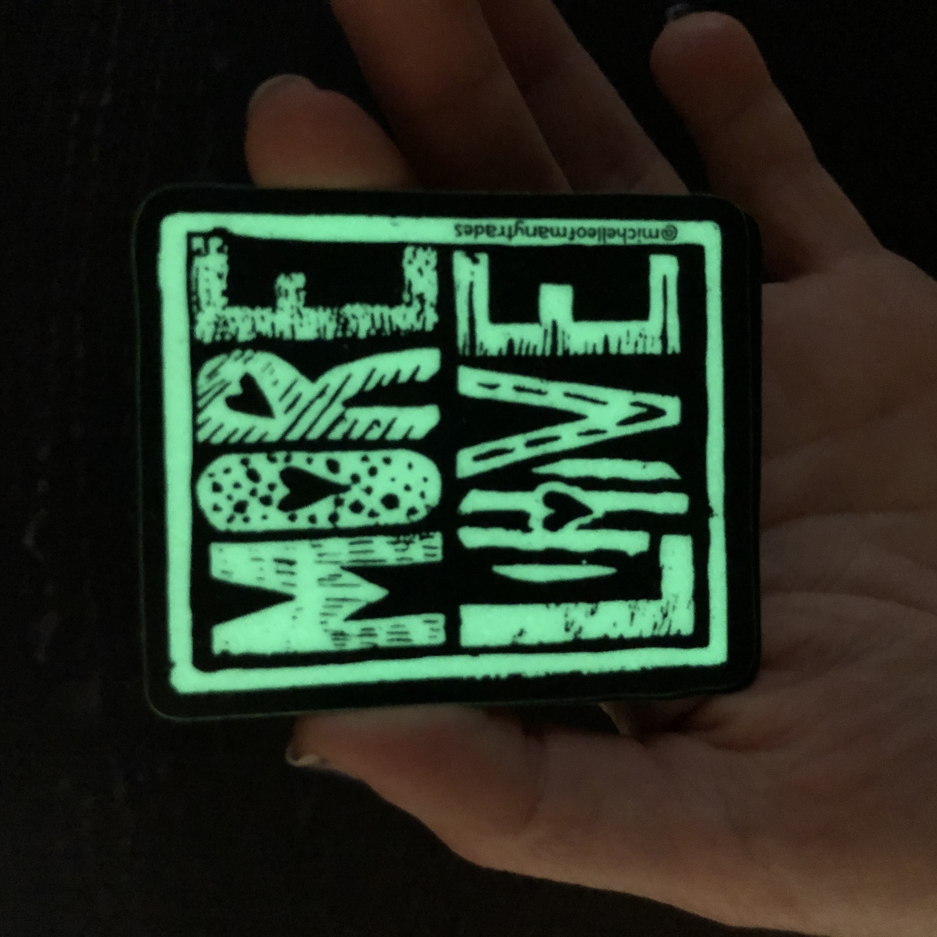 Michelle is holding a glow-in-the-dark More Love sticker in partial darkness and it is glowing. The hand is barely visible holding the sticker.