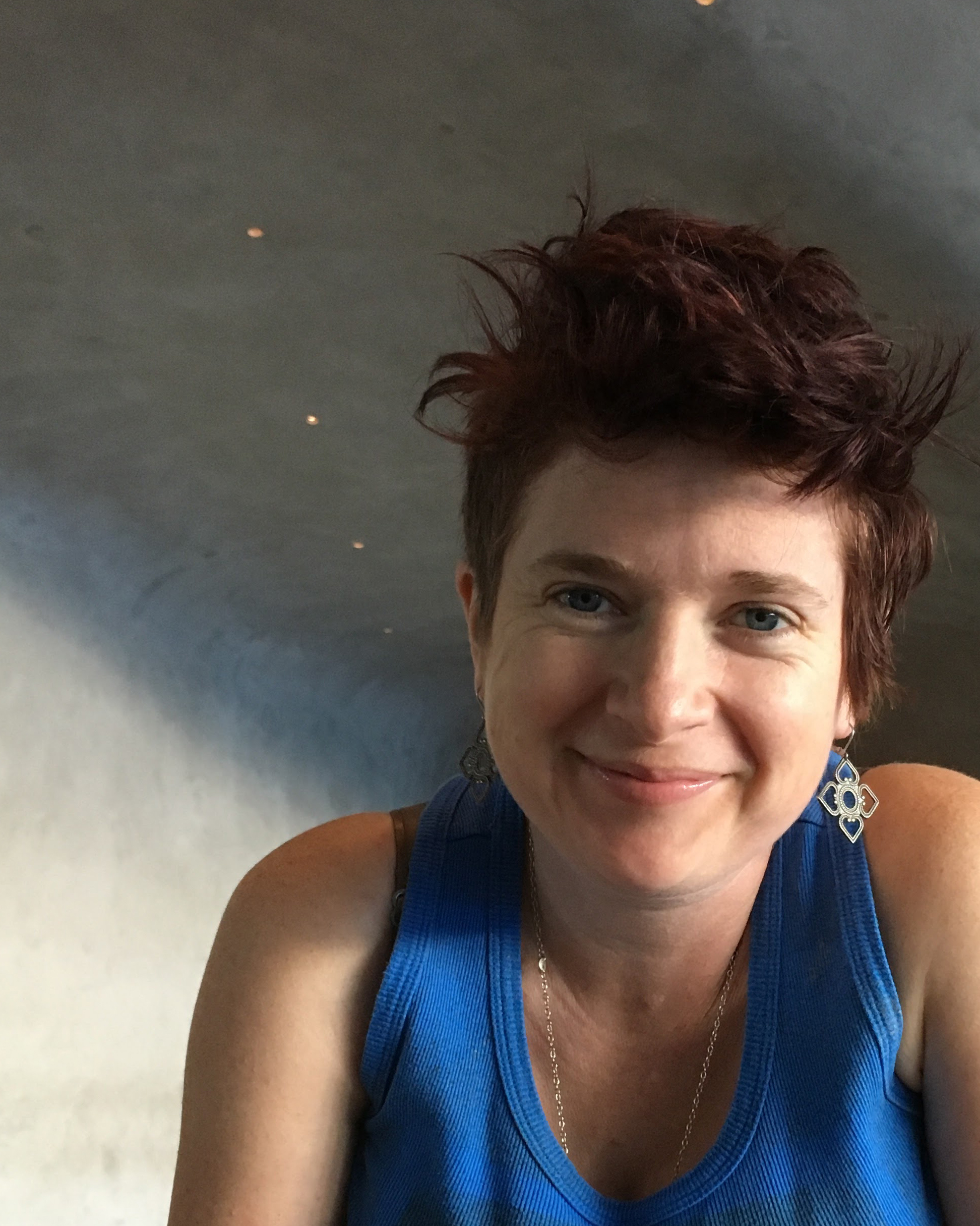 Michelle Rainville, a white nonbinary human, has a smile even in the eyes, shadow on part of their face, earrings in a metal flower shape, and is wearing a blue tank top. The concrete ceiling and wall are behind. There are pinpoints of light beyond.